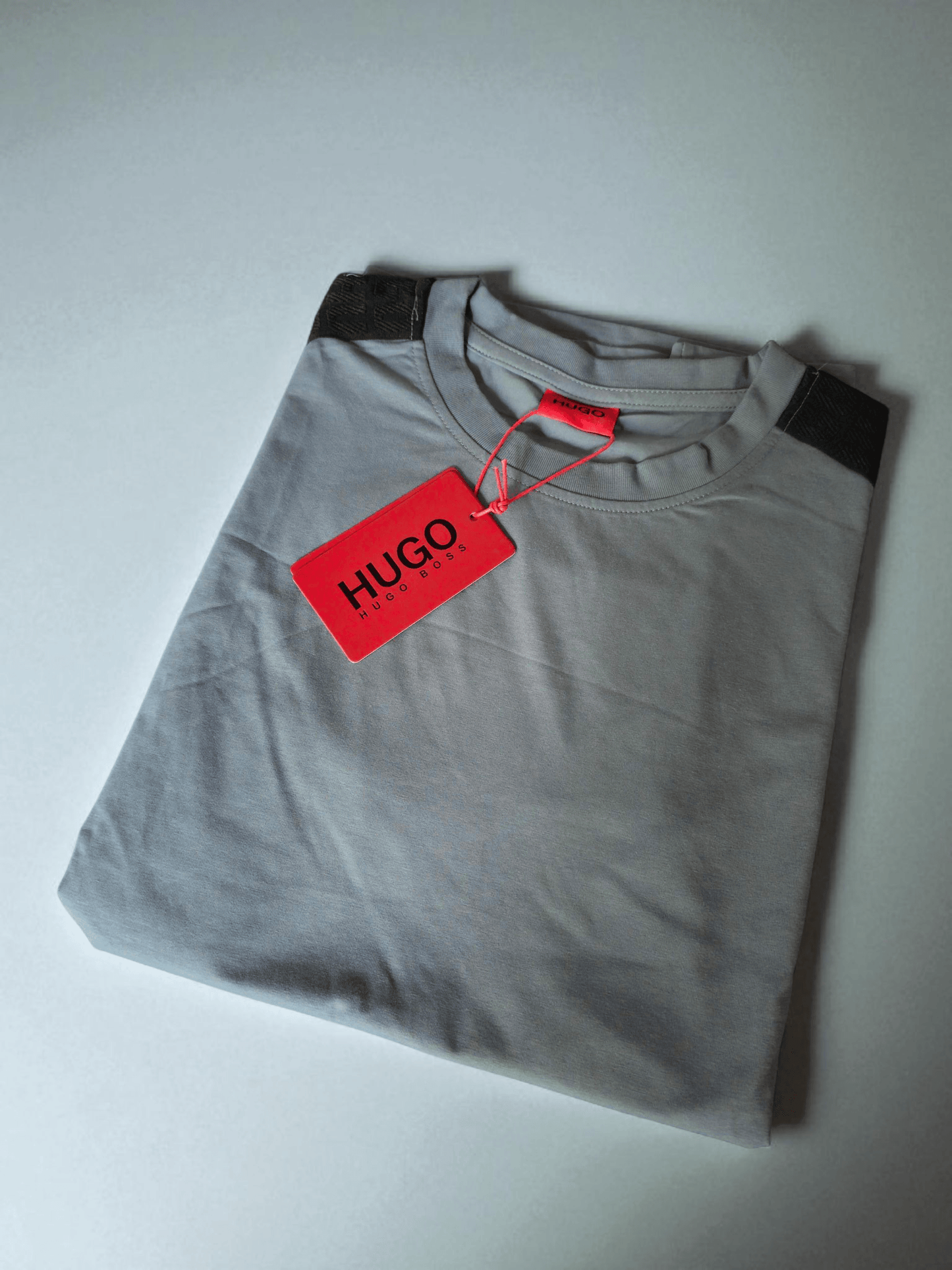Camiseta Hugo Boss gris claro doblada con etiqueta roja visible, confeccionada en algodón suave con detalles en los hombros. Estilo premium y cómodo.